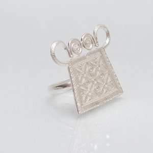Könnte beinhalten: Ein silberner Ring mit quadratischem Design, das mit komplizierten Details verziert ist. Der Ring hat eine einzigartige, verzierte Oberseite mit einem dekorativen Muster.