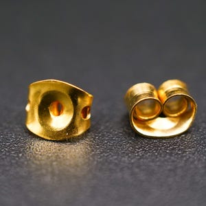 Peut inclure: Deux fermoirs de boucles d'oreilles dorés sur une surface sombre. L'un est un fermoir de type papillon avec un trou central et deux petits trous latéraux. L'autre est un fermoir de boucle d'oreille à double tube.