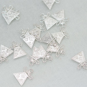 Puede incluir: Una colección de pequeños dijes de metal plateado. Cada dije presenta una forma triangular con un patrón floral detallado y detalles de alambre decorativo. Los dijes están dispersos sobre una superficie gris claro.