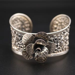 May include: A silver cuff bracelet with a textured band and a detailed flower design. The flower is the focal point, with multiple petals and a detailed center. The bracelet is set against a dark background.