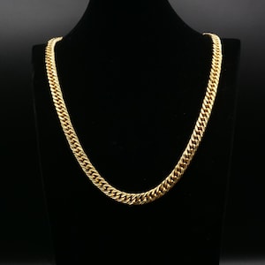 Puede incluir: Un collar de cadena de color dorado con un diseño de eslabones de acera. El collar se exhibe en un maniquí negro, mostrando su acabado pulido y su intrincado patrón. La cadena es un accesorio audaz.