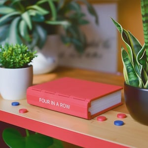 May include: A red game case labeled "FOUR IN A ROW" sits on a wooden shelf. The game case is rectangular and has a book-like design. Several red and blue game pieces are scattered around the case. Green plants in white and black pots are in the background.
