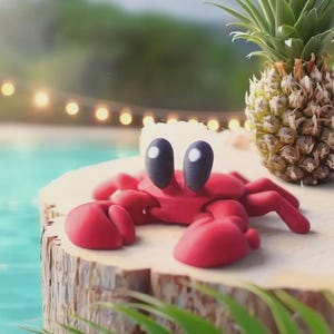 May include: A whimsical red crab figurine with large black eyes and claws rests on a wooden surface. A ripe pineapple with green leaves sits nearby. The scene evokes a tropical, summery vibe with a blurred pool and string lights in the background.