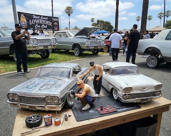 May include: A detailed display featuring model cars, including a silver lowrider and a white classic car, on a wooden table. Miniature figures are positioned around the cars, appearing to work on them. The scene is set outdoors with real cars and people in the background.