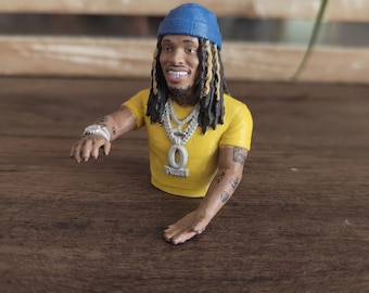 May include: A miniature figurine of a person wearing a blue beanie, a yellow t-shirt, and multiple silver chain necklaces with a large pendant. The figure has long dark dreadlocks with blonde highlights, tattoos on the arms, and a silver watch.