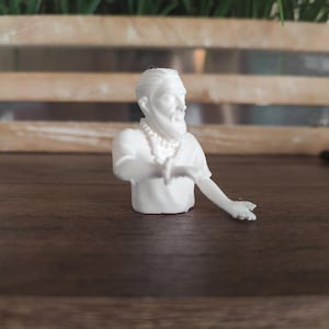 May include: A white 3D printed figurine of a person with a beard and hair tied back, wearing a short-sleeved shirt and a beaded necklace. The figure is depicted from the chest up with one arm extended outwards, showing detailed facial features.