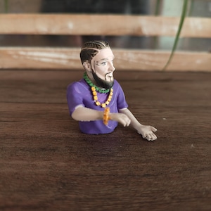 May include: A small figurine of a person with a beard and braided hair, wearing a purple t-shirt, a green beaded necklace with yellow accents, and a matching yellow beaded bracelet on the wrist. The figure is posed with one arm extended outwards.