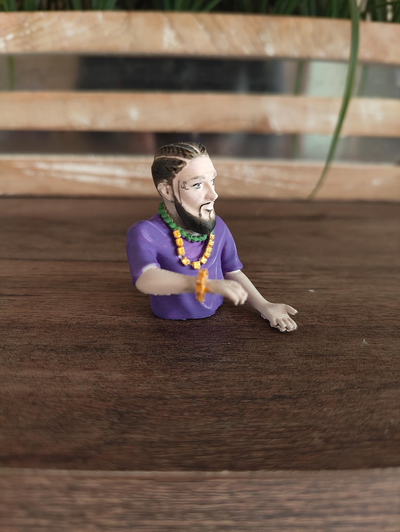 May include: A small figurine of a person with a beard and braided hair, wearing a purple t-shirt, a green beaded necklace with yellow accents, and a matching yellow beaded bracelet on the wrist. The figure is posed with one arm extended outwards.