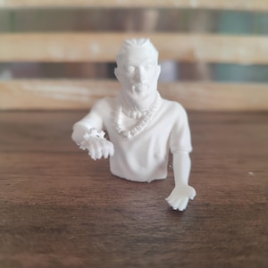May include: A white 3D printed bust of a person with braided hair, wearing a short-sleeved shirt, a beaded necklace, and a wrist accessory. The figure is depicted from the chest up with one arm extended forward and the other arm reaching out.