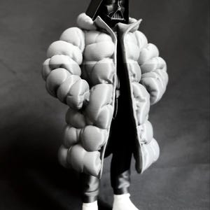 May include: A black and white image of a figure in a Darth Vader mask and helmet, wearing a puffy, light gray coat, black pants, and white boots. The coat has a large, quilted design. The figure is standing against a dark gray background.