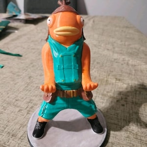 May include: A 3D printed figurine of a humanoid orange fish character with large eyes and thick lips. The figure wears a teal vest, teal cargo pants, a brown belt with pouches, a brown cap, and black shoes, standing on a circular base.