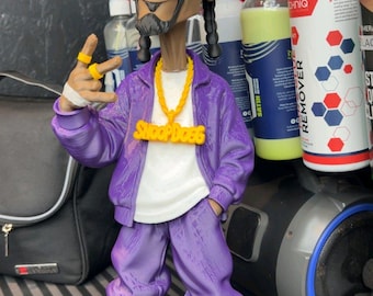 May include: A stylised figurine of a person with a goatee and braided hair, wearing a purple track jacket and matching trousers over a white t-shirt. The figure features a yellow chain necklace with the text SNOOP DOGG, yellow rings, and white trainers.