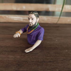 May include: A 3D-printed figurine of a person with braided hair, a dark beard, and a moustache. The figure wears a purple short-sleeved shirt, a green beaded necklace, an orange chain necklace, and an orange spiked bracelet on the right wrist.