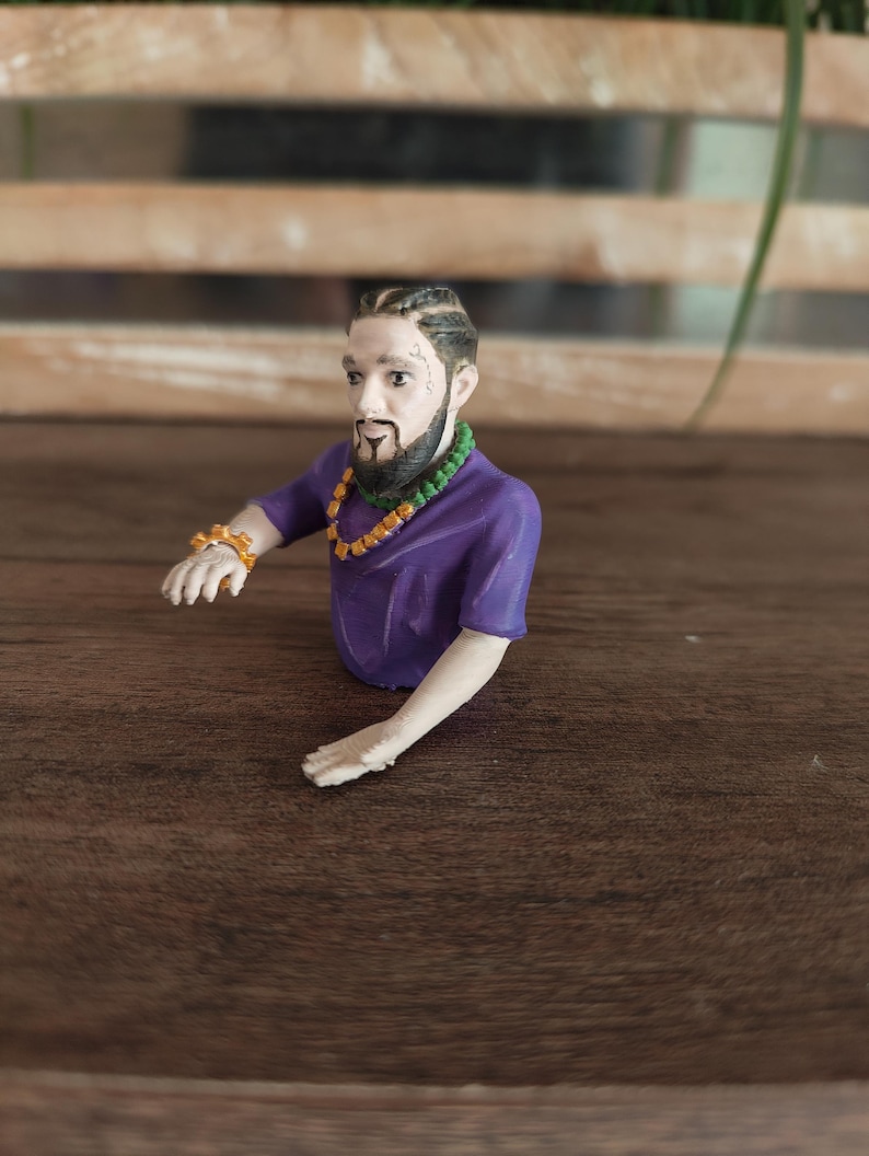 May include: A 3D-printed figurine of a person with braided hair, a dark beard, and a moustache. The figure wears a purple short-sleeved shirt, a green beaded necklace, an orange chain necklace, and an orange spiked bracelet on the right wrist.