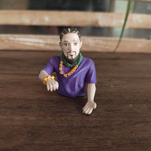May include: A small figurine of a person with a beard and braided hair, wearing a purple t-shirt, a green beaded necklace, a yellow square-beaded necklace, and a yellow beaded bracelet on the right wrist, with one hand extended forward.