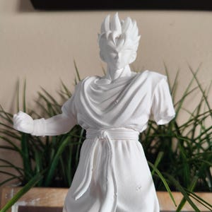 May include: A white figurine of a male anime character in a dynamic pose, with spiky hair and a flowing outfit. The figure is set against a backdrop of green plants in a wooden planter and a neutral-toned wall.