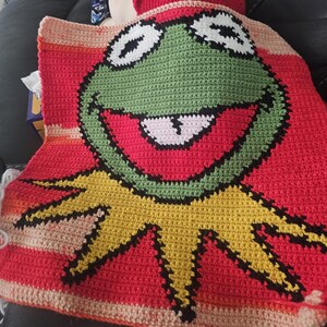 May include: A crocheted blanket featuring a large, smiling Kermit the Frog design. The blanket is primarily red with a yellow collar and green face. The frog's eyes are white with black outlines, and his mouth is open, revealing a pink tongue.