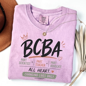 Op de afbeelding: Een lichtpaars t-shirt met korte mouwen met de tekst BCBA in grote zwarte letters en een kroonpictogram. Daaronder staat de tekst Part Scientist, Part Teacher, Part Advocate, All Heart en Changing Lives Daily in het groen en oranje.