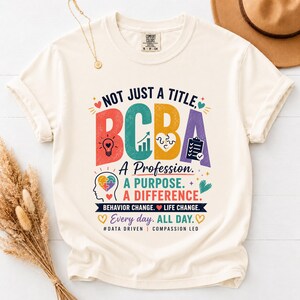 Könnte beinhalten: Ein cremefarbenes Kurzarm-T-Shirt mit einem mehrfarbigen Grafikdruck mit der Aufschrift: NOT JUST A TITLE, BCBA, A Profession. A PURPOSE. A DIFFERENCE. BEHAVIOR CHANGE. LIFE CHANGE. Every day. ALL DAY. #DATA DRIVEN | COMPASSION LED.