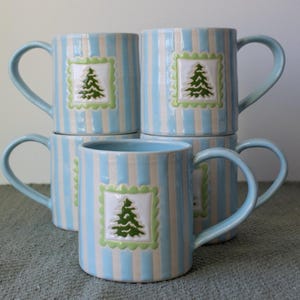 May include: A stack of four ceramic mugs with light blue and white vertical stripes. Each mug has a light blue handle and a square design with a green Christmas tree. The mugs are stacked on a gray surface.