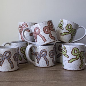 May include: A collection of handmade ceramic mugs with a speckled white finish. Each mug features a hand-painted bow design in shades of pink, green, and blue. The mugs have a curved handle and are stacked on a wooden surface.
