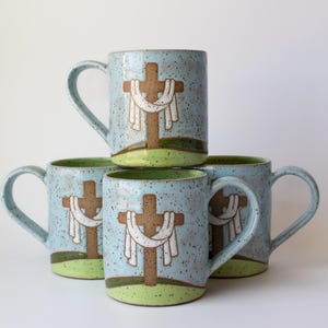 May include: Four ceramic mugs stacked, each with a light blue speckled finish. The mugs feature a brown cross design with a white cloth draped over it, set against a green base. The handles are light blue.