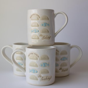 May include: Four white ceramic mugs stacked together. Each mug features a design of stylized fish and dumplings, with the text "Matthew 14:13-21" and "He Provides!" in a handwritten font. The fish are light blue.