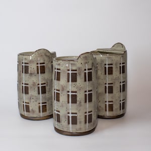 May include: Three ceramic vases with a speckled gray glaze and a repeating pattern of brown squares and white vertical lines. Each vase has a unique, curved top and a dark brown base.