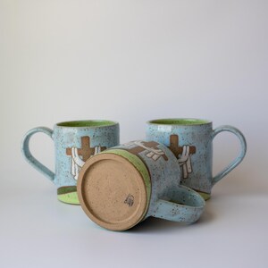 May include: Three ceramic mugs with a light blue speckled finish. Each mug features a brown cross design with white accents. The interior rim is green. One mug is lying on its side, showcasing the bottom.
