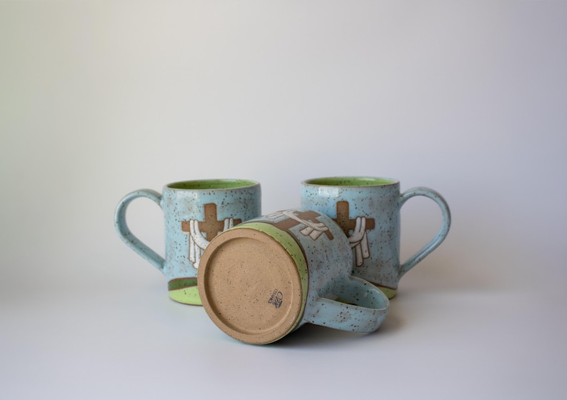May include: Three ceramic mugs with a light blue speckled finish. Each mug features a brown cross design with white accents. The interior rim is green. One mug is lying on its side, showcasing the bottom.