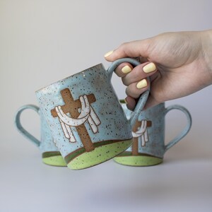 May include: Ceramic mugs with a light blue speckled finish, featuring a brown cross design with a white cloth. The mugs have a green base and a curved handle. The image shows three mugs, with one being held.