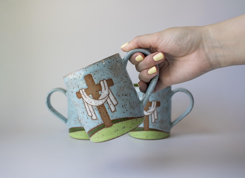 May include: Ceramic mugs with a light blue speckled finish, featuring a brown cross design with a white cloth. The mugs have a green base and a curved handle. The image shows three mugs, with one being held.