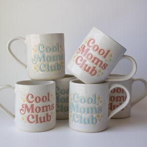 May include: Several white ceramic mugs, each bearing the text "Cool Moms Club" in pastel shades. The mugs are stacked, with the text in pink and blue, and have a curved handle, suitable for tea or coffee.
