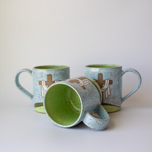 May include: Three ceramic mugs with a light blue speckled finish and a green interior. Each mug features a brown cross design with white accents. One mug is lying on its side. The mugs have curved handles.