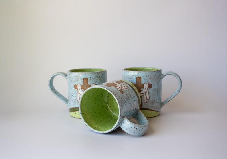 May include: Three ceramic mugs with a light blue speckled finish and a green interior. Each mug features a brown cross design with white accents. One mug is lying on its side. The mugs have curved handles.