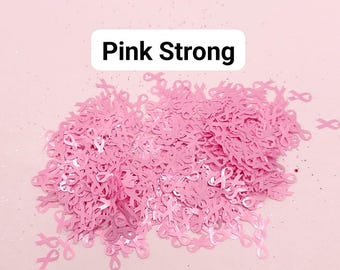 May include: Pink ribbon confetti, a symbol of breast cancer awareness, scattered on a pink background. The text "Pink Strong" is written on a white rectangle.