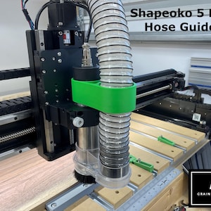 May include: A Shapeoko 5 Pro CNC machine with a green hose guide attached to a clear dust collection hose. The machine is on a wooden table with a ruler and a Crain Precision logo.