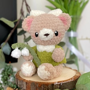 May include: A crocheted plush toy bear wearing a green dress and holding a sprig of white lily of the valley flowers. The bear has pink ears, white fur, and black eyes. The bear is sitting on a wooden slice.