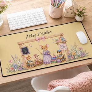 May include: A rectangular desk mat with a light yellow background featuring a whimsical illustration of mice in a classroom setting. The text Mrs Miller is printed at the top. The scene includes a teacher mouse, student mice, books, flowers, and a small chalkboard with the text Teach Love Inspire and a sign that says Be Kind Be Curious Be You.