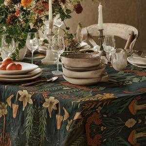 May include: A dining table set for a meal, featuring a dark teal tablecloth with a floral pattern. The table is set with white plates, glasses, and a centerpiece of flowers and candles. The room has a rustic, natural aesthetic.