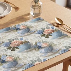 May include: A rectangular placemat featuring a repeating pattern of blue teacups filled with pink and blue flowers. The placemat is on a wooden table, with a matching napkin and gold utensils.