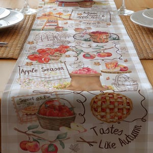 May include: A dining table set for a meal, featuring white dishes, wine glasses, and a decorative table runner. The runner has an autumn theme with illustrations of apples, pies, and text that reads "Apple Season" and "Tastes Like Autumn".