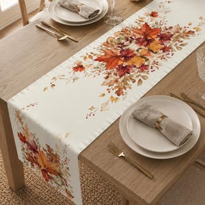 May include: A long, rectangular table runner featuring a cream-colored background with a vibrant, watercolor-style print of autumn leaves in shades of orange, red, yellow, and brown, arranged in clusters along the center and ends of the fabric.