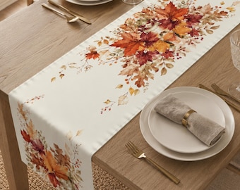 May include: A long, rectangular table runner featuring a cream-coloured background with a vibrant, watercolour-style print of autumn leaves in shades of orange, red, yellow, and brown, arranged in clusters along the centre and ends of the fabric.