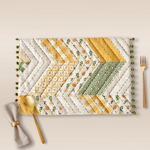 May include: A rectangular quilted placemat with a patchwork design in white, yellow, and green. The placemat features a chevron pattern with various floral and geometric prints. Green pom-poms trim the edges. A gold spoon, fork, and napkin with a ring are on the table.