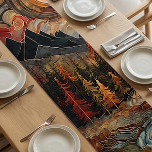 May include: A decorative table runner with a stained glass design featuring a mountain and forest scene in shades of black, brown, red, orange, and blue. The runner is placed on a light wood table, set with white plates, silverware, and napkins.