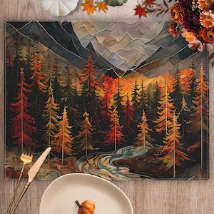 May include: Rectangular placemat featuring a forest scene with mountains, trees, and a river. The artwork uses warm colors like orange, red, and brown, evoking an autumn theme. A fork and knife are placed on the left, and a white plate with pumpkins is at the bottom.