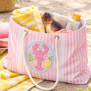 May include: Pink and white striped beach tote bag with a lobster patch. The bag is filled with a yellow and white striped towel, sunglasses, and sunscreen. The bag has white rope handles.