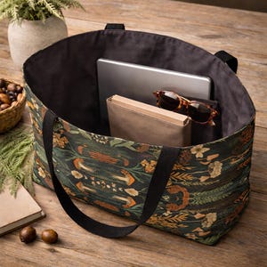 May include: A large tote bag with a dark floral pattern, black handles, and a black interior. The bag contains a tablet, sunglasses, and a book. The bag is sitting on a wooden surface.