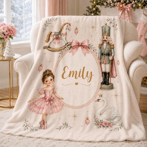 May include: A cream-colored fleece blanket featuring a central oval frame with the name Emily in gold script. The design includes a ballerina in a pink tutu, a rocking horse, a nutcracker soldier, and a white swan, accented with pink bows and stars.